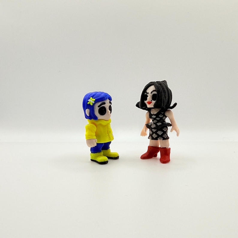 K&ouml;nnte beinhalten: Zwei kleine Figuren von Filmfiguren. Eine Figur hat blaue Haare, eine gelbe Jacke, blaue Hosen und gelbe Stiefel. Die andere Figur hat schwarze Haare, ein schwarz-wei&szlig;es Kleid und rote Stiefel.
