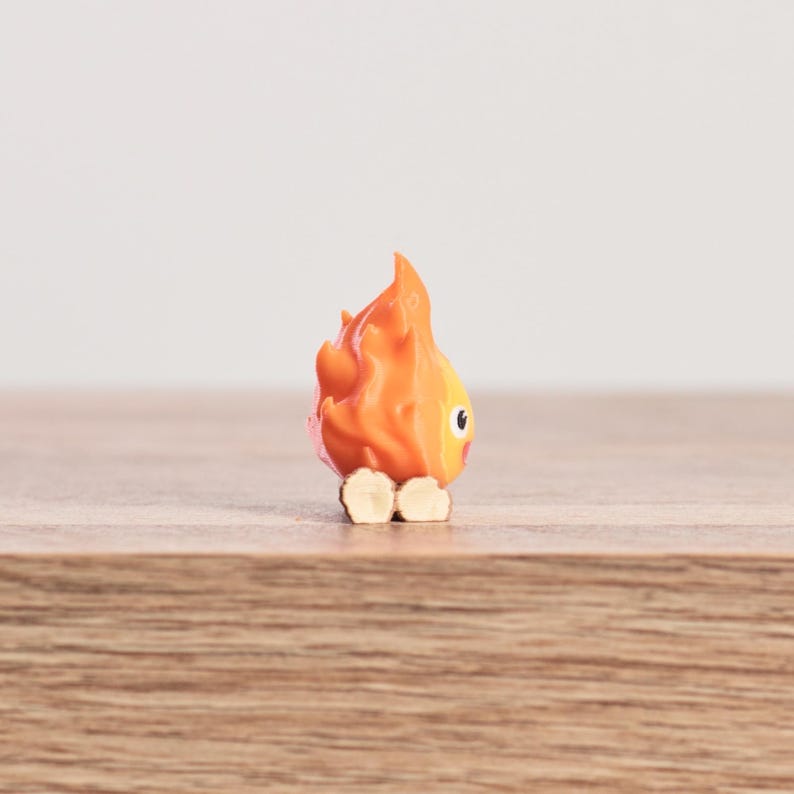 May include: A small, orange figurine shaped like a flame with a smiling face and small beige feet. The figurine is set against a light background and sits on a wooden surface. The flame has a cartoonish design.