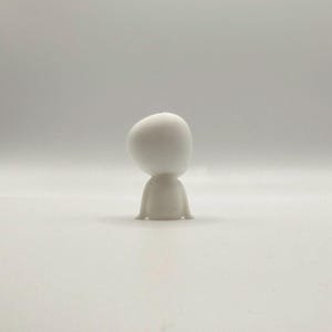 Op de afbeelding: Een klein, wit beeldje van een gestileerde persoon, van achteren bekeken. De figuur heeft een afgeronde kop en een eenvoudig lichaam met kleine, uitlopende voeten. Het beeldje staat tegen een effen, lichte achtergrond.