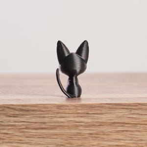 May include: A small, black cat figurine. The cat is depicted looking over its shoulder, with its tail curved upwards. The figurine is made of a solid, matte black material and sits on a wooden surface. The cat's ears are pointed.