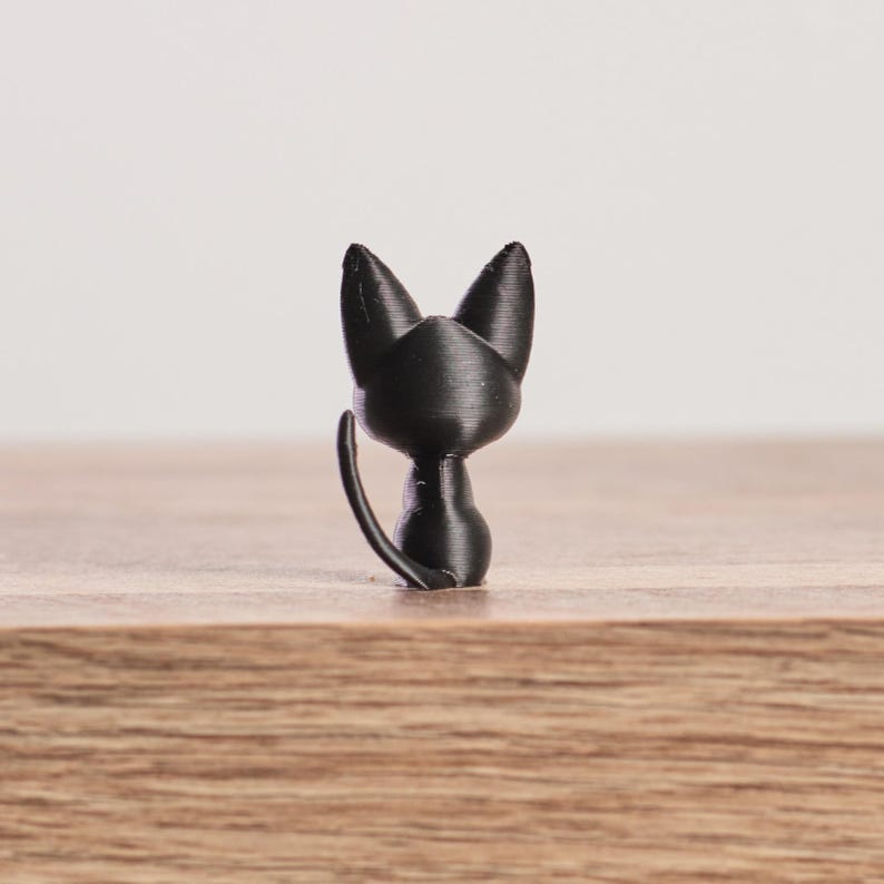 May include: A small, black cat figurine. The cat is depicted looking over its shoulder, with its tail curved upwards. The figurine is made of a solid, matte black material and sits on a wooden surface. The cat's ears are pointed.