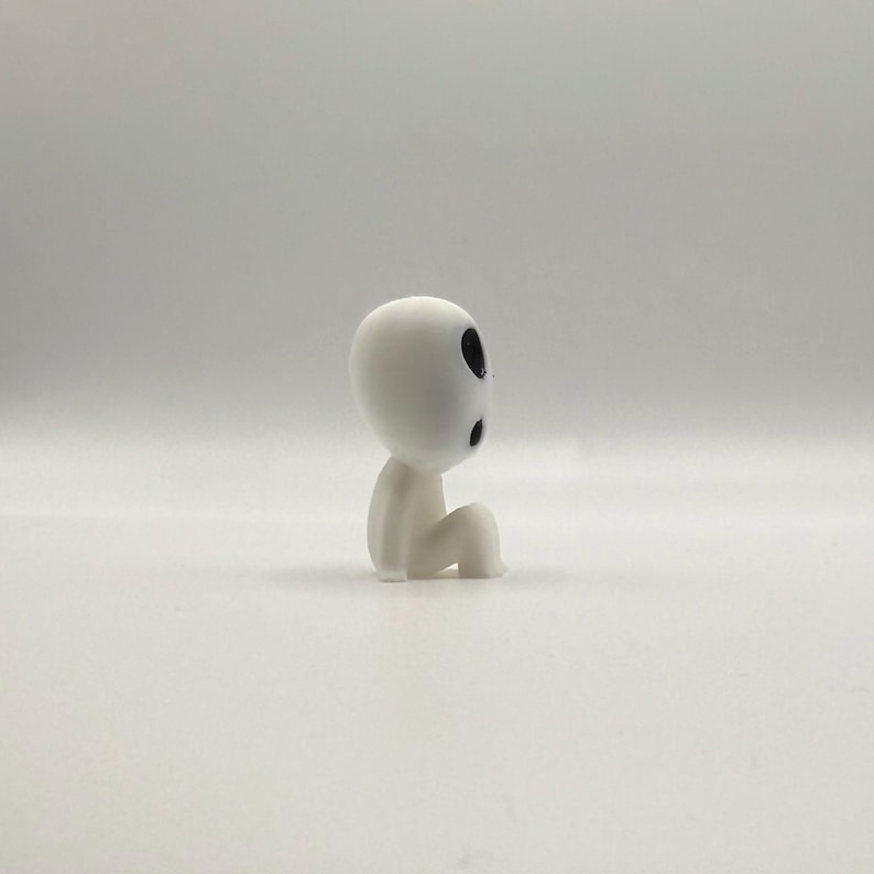 Op de afbeelding: Een klein, wit beeldje van een zittende bosgeest met zwarte oogmarkeringen. De figuur heeft een gladde, afgeronde kop en een eenvoudig, minimalistisch ontwerp, dat een gevoel van rust en fantasie oproept. Een verzamelobject.