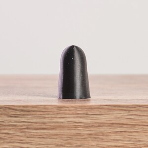 K&ouml;nnte beinhalten: Ein schwarzer, zylindrischer Flaschenverschluss mit abgerundeter Spitze. Der Verschluss besteht aus einem festen Material und hat eine glatte, matte Oberfl&auml;che. Er steht auf einer Holzoberfl&auml;che.