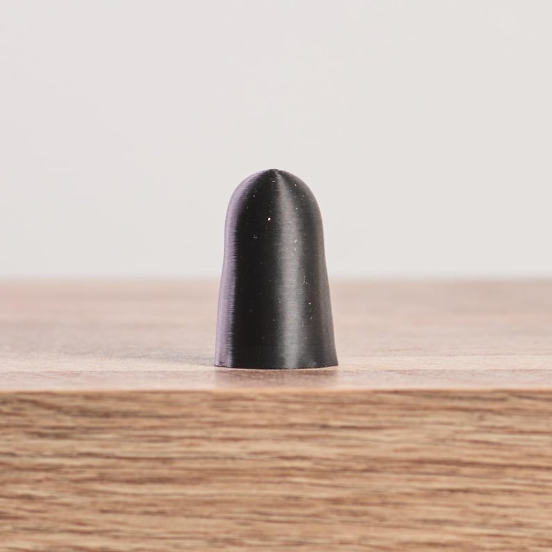 K&ouml;nnte beinhalten: Ein schwarzer, zylindrischer Flaschenverschluss mit abgerundeter Spitze. Der Verschluss besteht aus einem festen Material und hat eine glatte, matte Oberfl&auml;che. Er steht auf einer Holzoberfl&auml;che.