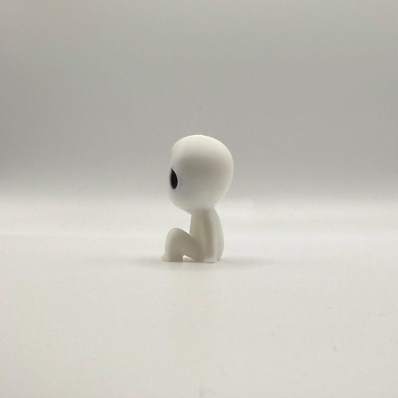 Op de afbeelding: Een klein, wit beeldje van een zittende humano&iuml;de figuur met een ronde kop en grote, zwarte ogen. De figuur zit in een zittende positie, met gebogen knie&euml;n en armen die op de benen rusten. Het beeldje staat op een witte ondergrond.