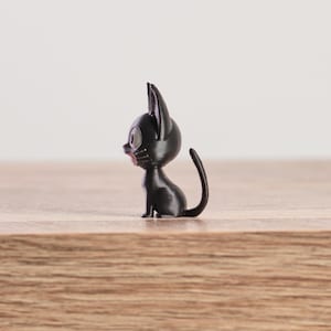 May include: A small, black cat figurine with large, round eyes and pointed ears. The cat is sitting upright with its tail curved. The figurine is on a wooden surface.