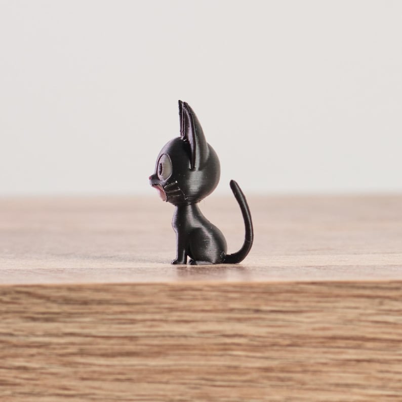 May include: A small, black cat figurine with large, round eyes and pointed ears. The cat is sitting upright with its tail curved. The figurine is on a wooden surface.