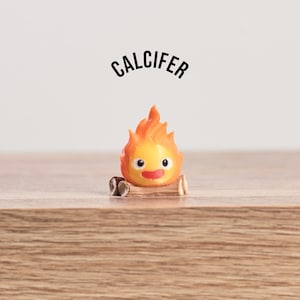 May include: A small, orange and yellow figurine of Calcifer, a fire demon, with a smiling face and flames. The figurine is perched on a small log, with the name "CALCIFER" arched above it in black text.