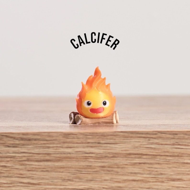 May include: A small, orange and yellow figurine of Calcifer, a fire demon, with a smiling face and flames. The figurine is perched on a small log, with the name "CALCIFER" arched above it in black text.