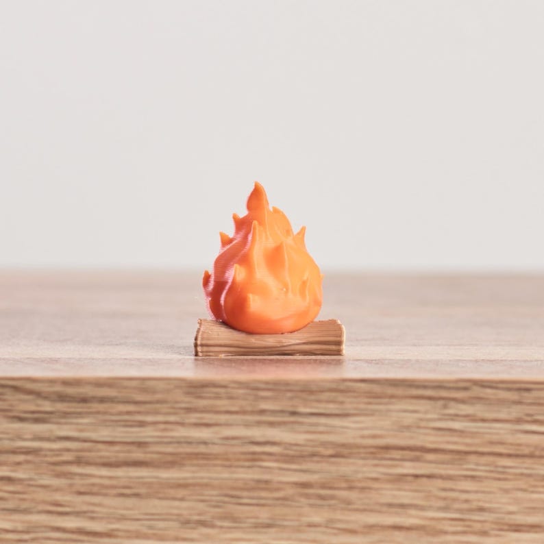 May include: A miniature orange flame sculpture atop a small, brown log. The flame is a vibrant orange color, with detailed peaks and valleys. The log appears to be made of wood, with visible grain. The sculpture is likely a decorative item or a toy.