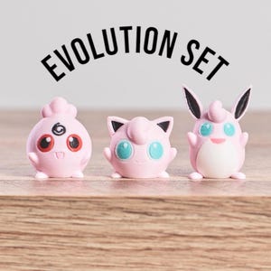 May include: A set of three pink figurines, each with unique features, standing on a wooden surface. The figurines have black accents and large eyes. The text "EVOLUTION SET" is arched above the figurines.