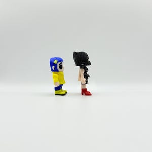 K&ouml;nnte beinhalten: Zwei kleine Figuren von Charakteren aus einem Film. Eine Figur tr&auml;gt eine gelbe Jacke, blaue Hose und einen blauen Hut mit einer Blume. Die andere Figur tr&auml;gt ein schwarzes Kleid und rote Stiefel. Beide Figuren stehen nebeneinander.