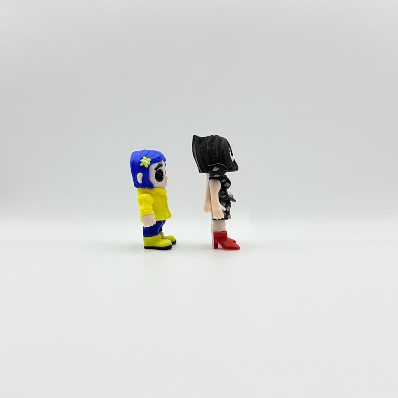 K&ouml;nnte beinhalten: Zwei kleine Figuren von Charakteren aus einem Film. Eine Figur tr&auml;gt eine gelbe Jacke, blaue Hose und einen blauen Hut mit einer Blume. Die andere Figur tr&auml;gt ein schwarzes Kleid und rote Stiefel. Beide Figuren stehen nebeneinander.