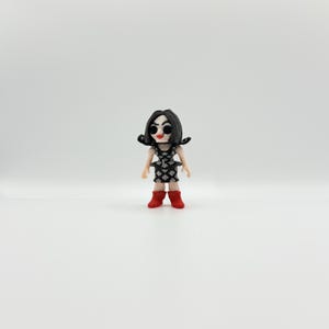 May include: A small doll with black hair, red boots, and a black and white patterned dress. The doll has a pale complexion, red lips, and large black eyes. The doll is standing on a white surface.