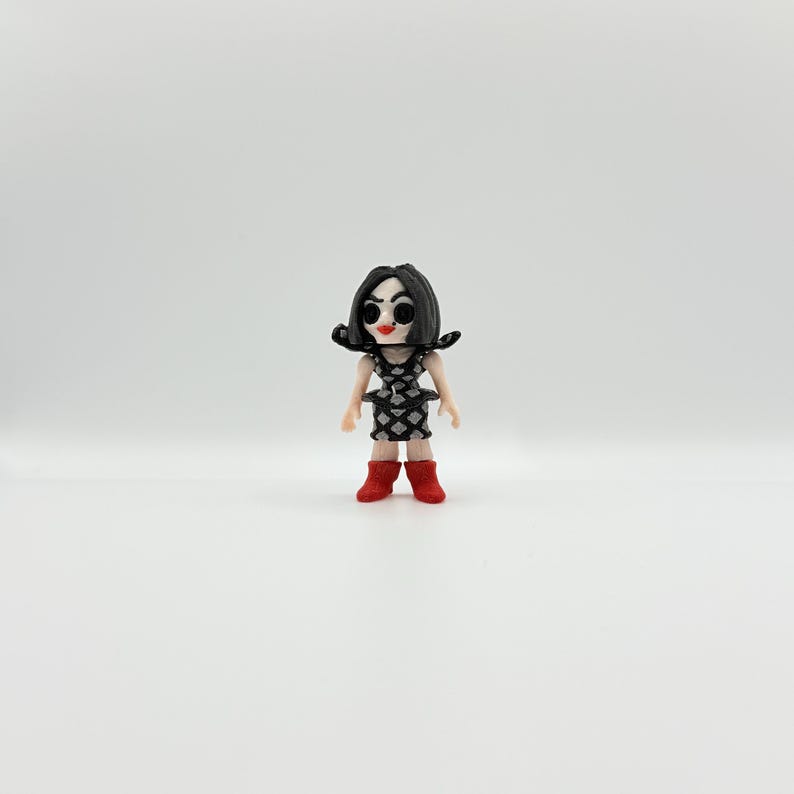 May include: A small doll with black hair, red boots, and a black and white patterned dress. The doll has a pale complexion, red lips, and large black eyes. The doll is standing on a white surface.