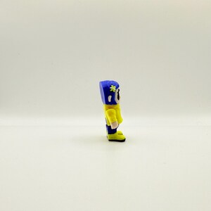 May include: A small, handmade figurine of a character in a yellow jacket, blue pants, and a blue helmet with a flower. The figurine has a cartoonish design and is standing on a white surface.