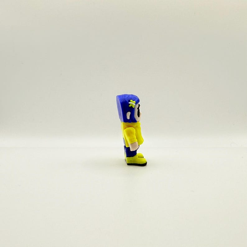 May include: A small, handmade figurine of a character in a yellow jacket, blue pants, and a blue helmet with a flower. The figurine has a cartoonish design and is standing on a white surface.