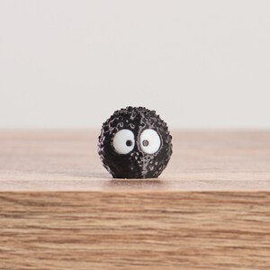 May include: A small, round, black figurine with a textured surface and large, white eyes. The figurine is set against a light background and rests on a wooden surface. The design is reminiscent of a cartoon character.