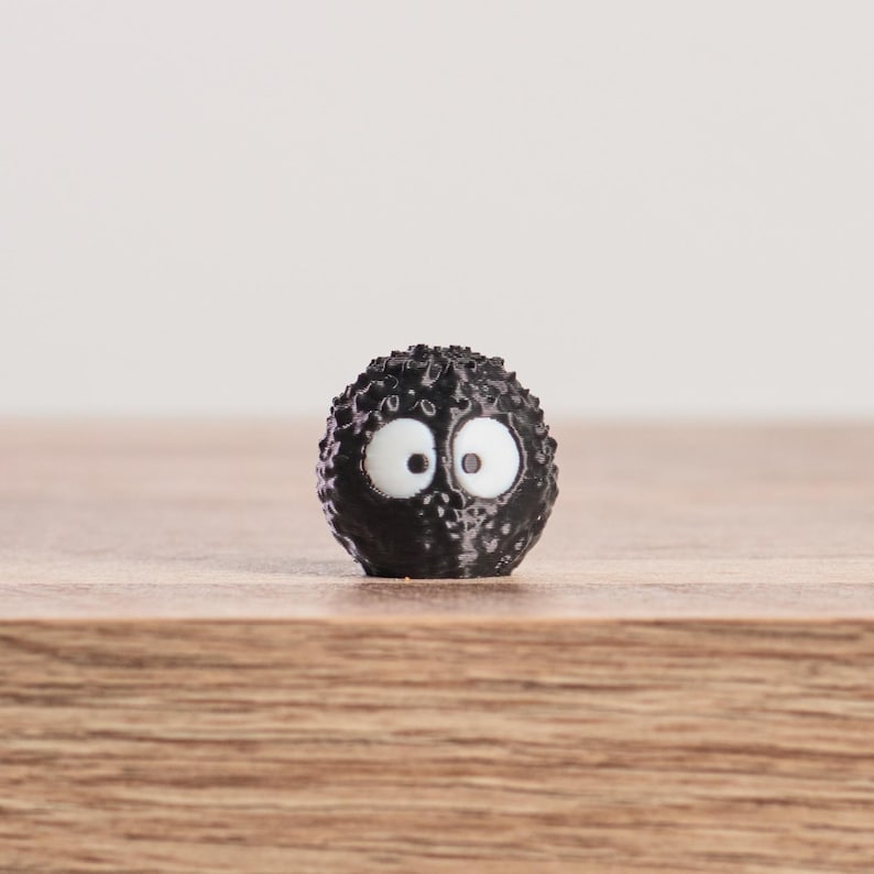 May include: A small, round, black figurine with a textured surface and large, white eyes. The figurine is set against a light background and rests on a wooden surface. The design is reminiscent of a cartoon character.