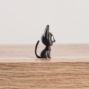 May include: A small, black cat figurine with large, round eyes and a pink mouth. The cat is sitting upright with its tail curled. The figurine is on a wooden surface.