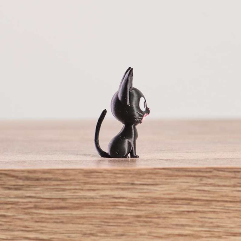 May include: A small, black cat figurine with large, round eyes and a pink mouth. The cat is sitting upright with its tail curled. The figurine is on a wooden surface.