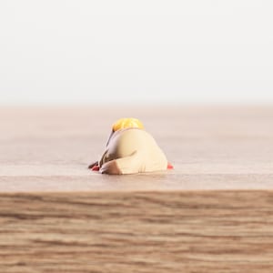 K&ouml;nnte beinhalten: Kleine, beige Figur einer sich vorbeugenden Person mit gelbem Hut und roten Akzenten. Die Figur liegt auf einer Holzoberfl&auml;che mit verschwommenem wei&szlig;em Hintergrund.