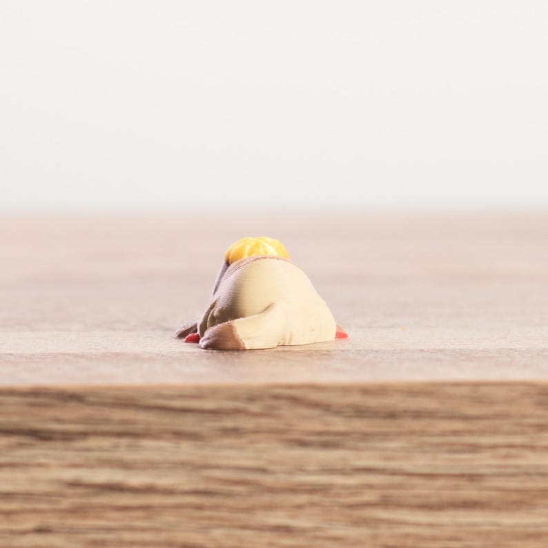 K&ouml;nnte beinhalten: Kleine, beige Figur einer sich vorbeugenden Person mit gelbem Hut und roten Akzenten. Die Figur liegt auf einer Holzoberfl&auml;che mit verschwommenem wei&szlig;em Hintergrund.
