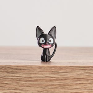 May include: A black cat figurine with large, pointed ears, wide white eyes, and a pink mouth. The cat is sitting upright with its tail curved. The figurine is on a wooden surface.