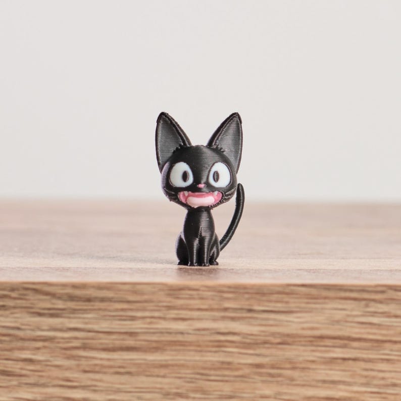 May include: A black cat figurine with large, pointed ears, wide white eyes, and a pink mouth. The cat is sitting upright with its tail curved. The figurine is on a wooden surface.