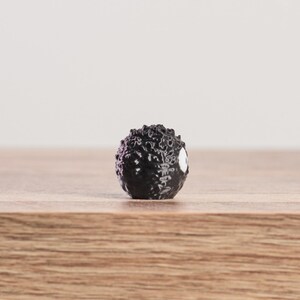 May include: A small, black, spherical figurine with a textured surface and a white eye-like marking. The figurine is set against a light background and rests on a wooden surface. The object appears to be a miniature sculpture or collectible.