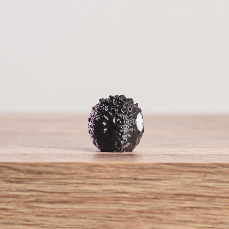 May include: A small, black, spherical figurine with a textured surface and a white eye-like marking. The figurine is set against a light background and rests on a wooden surface. The object appears to be a miniature sculpture or collectible.