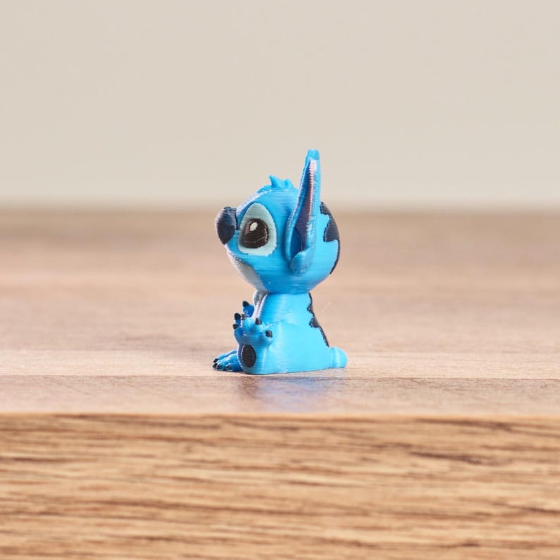 Peut inclure: Petite figurine bleue clair du personnage Disney Stitch. La figurine a de grands yeux expressifs, des taches noires et de grandes oreilles. Elle est pos&eacute;e sur une surface en bois.