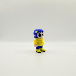 May include: A small figurine of a character with blue hair, a yellow jacket, blue pants, and yellow boots. The character has a flower in their hair and large black eyes. The figurine is standing on a white surface.