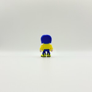 May include: A small, stylized figurine of a character from the animated film, "Coraline." The figure is viewed from the back, showcasing a blue bob hairstyle, a yellow jacket, blue pants, and yellow shoes with black accents. The background is plain white.