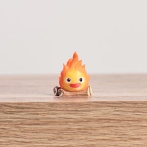 May include: A small, orange and yellow flame character figurine with a smiling face and a log base. The figurine has a cartoonish design with a bright, cheerful appearance. The figurine is a collectible item.