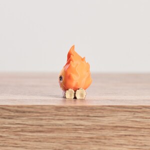 May include: A small, orange and yellow figurine of a flame with a face. The figurine has a cartoonish design, with a round body and a flame-shaped head. The figurine is sitting on a wooden surface.