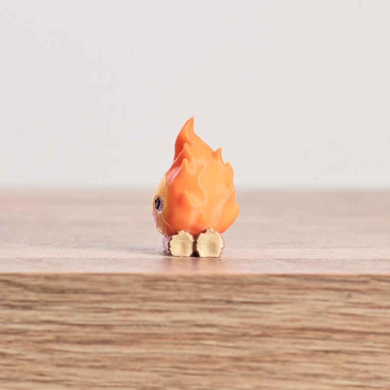 May include: A small, orange and yellow figurine of a flame with a face. The figurine has a cartoonish design, with a round body and a flame-shaped head. The figurine is sitting on a wooden surface.