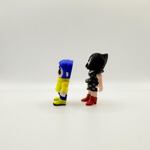 K&ouml;nnte beinhalten: Zwei kleine Figuren von Charakteren aus dem Animationsfilm "Coraline". Eine Figur tr&auml;gt eine gelbe Jacke, blaue Haare und blaue Stiefel. Die andere Figur tr&auml;gt ein schwarzes Kleid und rote Stiefel. Die Figuren stehen auf einer wei&szlig;en Oberfl&auml;che.