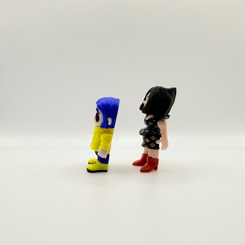 K&ouml;nnte beinhalten: Zwei kleine Figuren von Charakteren aus dem Animationsfilm "Coraline". Eine Figur tr&auml;gt eine gelbe Jacke, blaue Haare und blaue Stiefel. Die andere Figur tr&auml;gt ein schwarzes Kleid und rote Stiefel. Die Figuren stehen auf einer wei&szlig;en Oberfl&auml;che.