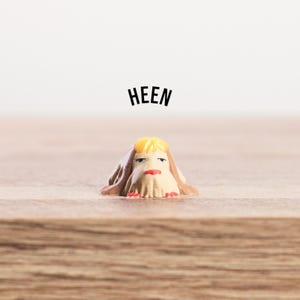 K&ouml;nnte beinhalten: Eine kleine, skurrile Figur mit beigem K&ouml;rper, gelben Haaren und rotem Mund. Die Figur ist mit einem beigen Tuch drapiert und tr&auml;gt das Wort "HEEN" in schwarzer Schrift dar&uuml;ber. Auf einer Holzoberfl&auml;che platziert.