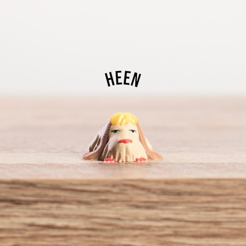 K&ouml;nnte beinhalten: Eine kleine, skurrile Figur mit beigem K&ouml;rper, gelben Haaren und rotem Mund. Die Figur ist mit einem beigen Tuch drapiert und tr&auml;gt das Wort "HEEN" in schwarzer Schrift dar&uuml;ber. Auf einer Holzoberfl&auml;che platziert.