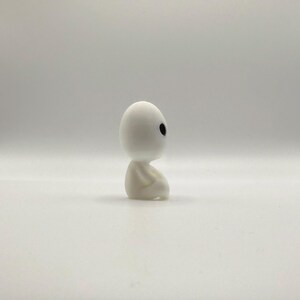 May include: A small, white figurine of a seated forest spirit with a rounded head and black eyes. The figure is in a seated position with its hands clasped. The figurine is on a white background.
