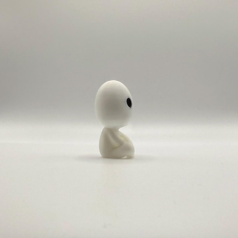May include: A small, white figurine of a seated forest spirit with a rounded head and black eyes. The figure is in a seated position with its hands clasped. The figurine is on a white background.