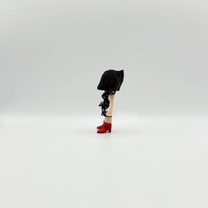 May include: A quirky figurine of a person with a black skirt and head covering. The figure's legs are visible, wearing a black and white patterned top and red heeled boots. The background is plain white.