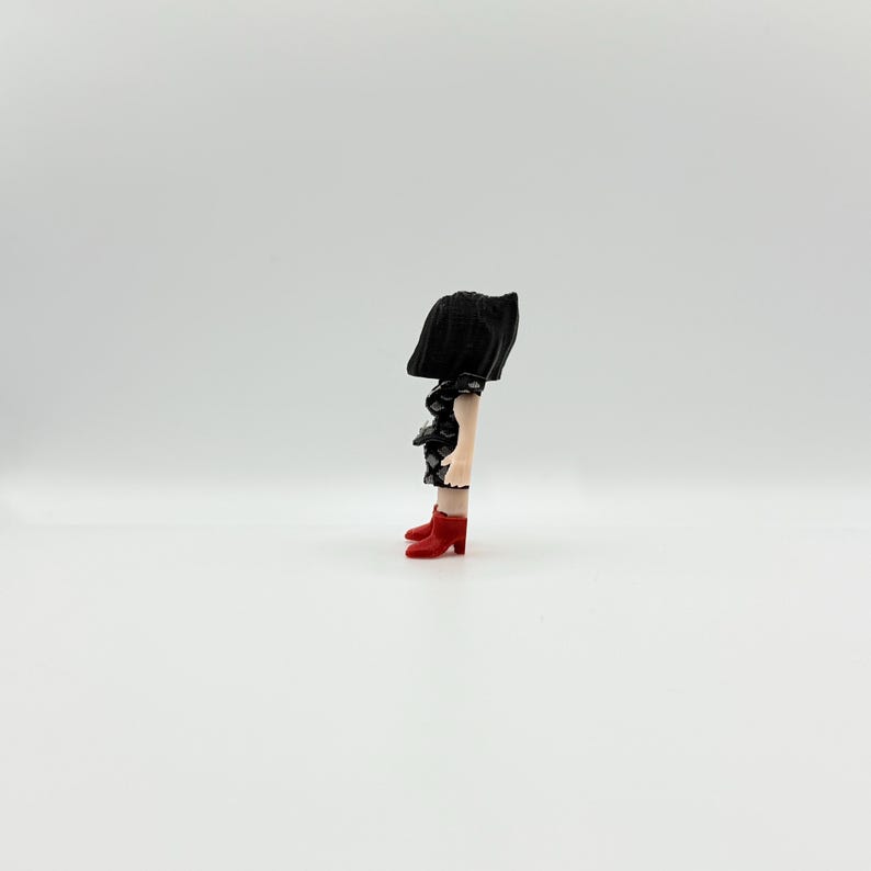 May include: A quirky figurine of a person with a black skirt and head covering. The figure's legs are visible, wearing a black and white patterned top and red heeled boots. The background is plain white.
