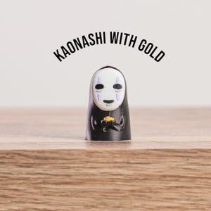 K&ouml;nnte beinhalten: Eine schwarze Kaonashi-Figur, eine Figur aus Spirited Away, die Gold h&auml;lt. Die Figur hat eine wei&szlig;e Maske mit lila Markierungen und einen schwarzen Umhang. Der Text "KAONASHI WITH GOLD" ist dar&uuml;ber gew&ouml;lbt.