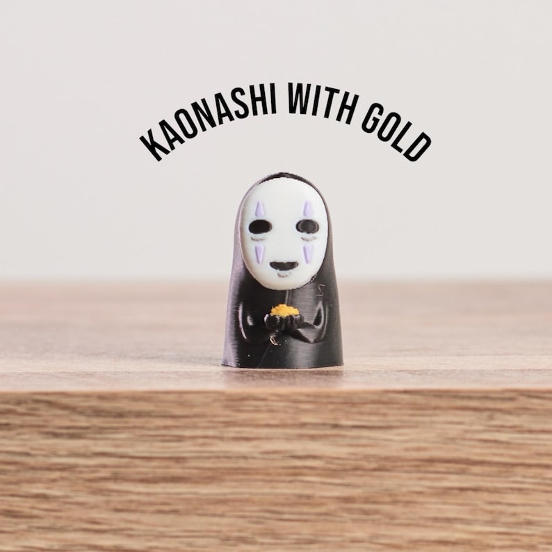 K&ouml;nnte beinhalten: Eine schwarze Kaonashi-Figur, eine Figur aus Spirited Away, die Gold h&auml;lt. Die Figur hat eine wei&szlig;e Maske mit lila Markierungen und einen schwarzen Umhang. Der Text "KAONASHI WITH GOLD" ist dar&uuml;ber gew&ouml;lbt.