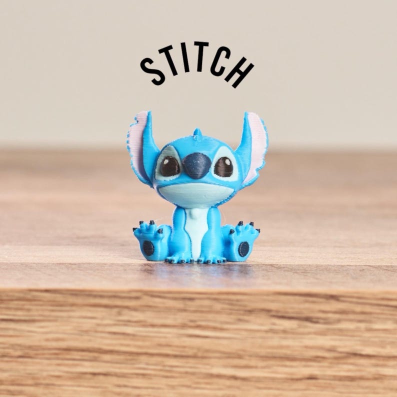 Peut inclure: Figurine bleue de Stitch avec de grandes oreilles, assis sur une surface en bois. La figurine a des yeux noirs et un ventre bleu clair. Le mot "STITCH" est &eacute;crit au-dessus de la figurine en noir, en texte incurv&eacute;.