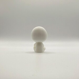 May include: A small, white figurine of a seated figure with a large, rounded head. The figure is in a meditative pose, with its legs crossed and arms resting on its knees. The figurine is made of a smooth, matte material.