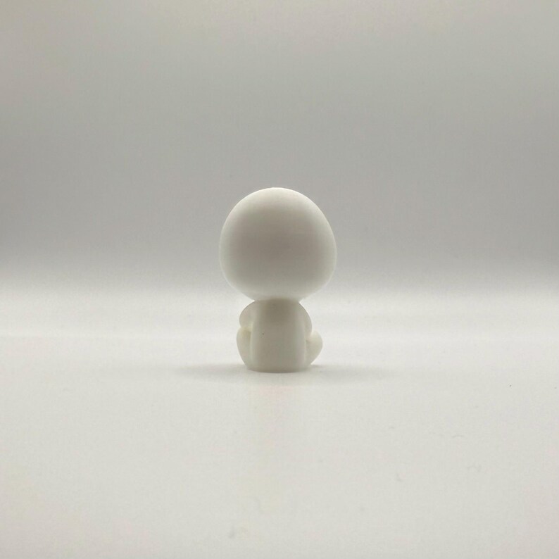 May include: A small, white figurine of a seated figure with a large, rounded head. The figure is in a meditative pose, with its legs crossed and arms resting on its knees. The figurine is made of a smooth, matte material.